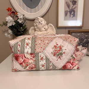 Puede incluir: Un neceser acolchado con un diseño de patchwork. La bolsa presenta una variedad de estampados florales en tonos rosa, rojo y verde. Un asa está unida al lateral. La bolsa está sobre una superficie blanca.