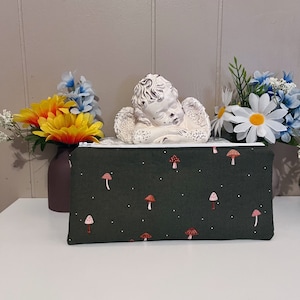 May include: A dark green zippered pouch with a white zipper and a repeating pattern of red and pink mushrooms. The pouch is sitting on a white surface in front of a sleeping cherub figurine and flowers.