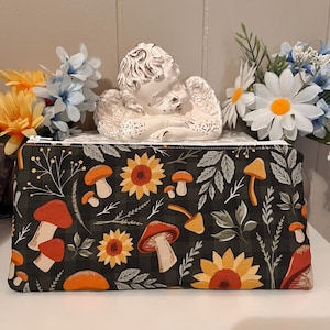 May include: A black zippered pouch with a floral pattern featuring mushrooms, sunflowers, and leaves. The pouch is on a white surface with a white ceramic angel figurine in the background.