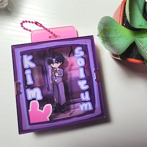 May include: A square purple acrylic frame featuring a central illustration of a cartoon character in a suit. The text KIM SOLTUM is printed in light purple on the sides. A pink plastic attachment with a pink ball chain is at the top edge.