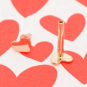 May include: Two gold-colored heart-shaped body jewelry pieces. One is a small stud, and the other has a long post. The jewelry is displayed on a white surface with red heart patterns.