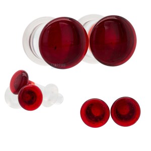 May include: A set of circular body jewelry plugs featuring a deep red, translucent glass front face and a clear, cylindrical stem. The design includes a smooth, rounded surface on the front and a flared back, with the text Starfire Body Jewelry visible.