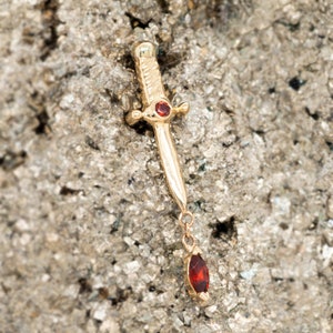 May include: A gold-colored pendant in the shape of a small dagger, featuring a red gemstone at the hilt and a dangling, teardrop-shaped red gemstone. The pendant is set against a textured, gray background.