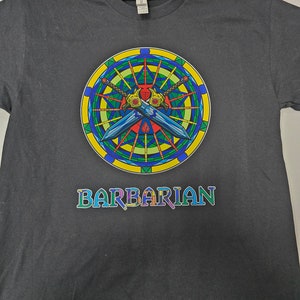 May include: A dark gray t-shirt with a colorful stained glass design featuring two crossed swords. The design is surrounded by a yellow circle with a blue and green border. The word "BARBARIAN" is printed in colorful letters below the design.