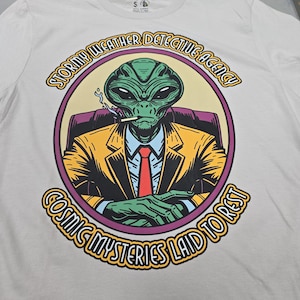 May include: A white t-shirt with a graphic of a green alien wearing a yellow suit and red tie. The alien is smoking a cigarette and sitting in a chair. The text around the alien reads "Stormy Weather Detective Agency Cosmic Mysteries Laid to Rest".