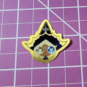 May include: A gold-colored sticker with a cartoon character design. The character has a black hat, dark blue hair, and large eyes with a heart and star design. The sticker is on a purple cutting mat.