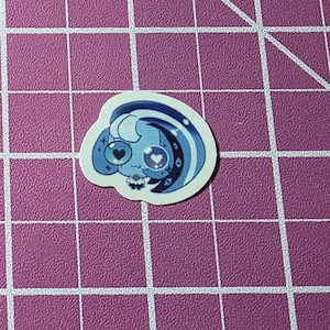 May include: A die-cut sticker featuring a blue and white cartoon character with heart-shaped eyes and a crescent moon design. The sticker has a white border and is set against a pink grid background.