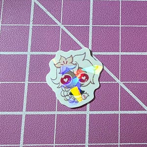 Può includere: Un piccolo adesivo olografico raffigurante un personaggio dei cartoni animati con accenti viola, rossi e gialli. Il personaggio ha grandi occhi e un design stravagante. L'adesivo è su un tappetino da taglio viola con un motivo a griglia.