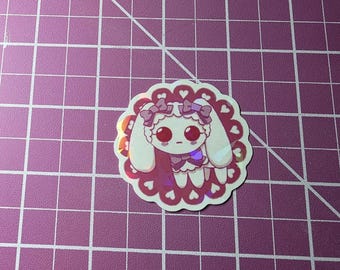 May include: A holographic sticker featuring a cartoon bunny with large eyes, wearing a bow and a dress. The sticker has a scalloped edge with heart shapes and is set against a purple grid background.