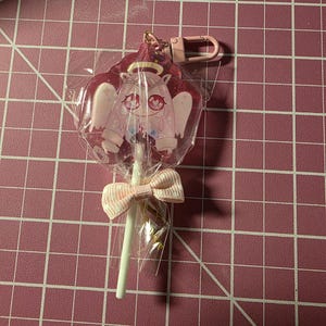 Puede incluir: Un llavero rosa con temática de ángel. El dije está en un envoltorio de plástico transparente y presenta un diseño de ángel de dibujos animados con un halo y alas. Un lazo rosa está atado alrededor del palo. Un cierre rosa está adjunto en la parte superior.