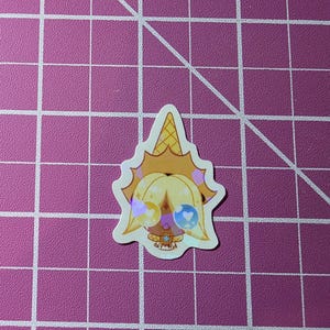 May include: A die-cut sticker featuring a cartoon character with a golden ice cream cone hat and large, round eyes. The character has yellow hair and wears a necklace. The sticker has a holographic effect and is set against a pink grid background.