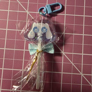 May include: A clear plastic-wrapped bunny-shaped keychain with a light blue clasp. The bunny has white ears, blue heart-shaped eyes, and a purple outline. A light blue bow is tied around the stick with gold string.