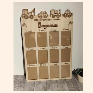 Può includere: Una cornice per foto in legno con 12 spazi per foto, ognuno etichettato con un mese. La cornice è decorata con un tema automobilistico e il testo "Ma première année" e "Benjamin".