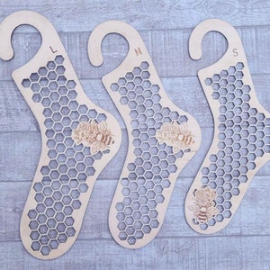 Op de afbeelding: Drie houten sokkenhangers met een honingraatpatroon en bijen- en bloemmotieven. De hangers zijn gemarkeerd met L, M en S. De hangers zijn gemaakt van lichtgekleurd hout en hebben een gebogen bovenkant om op te hangen.