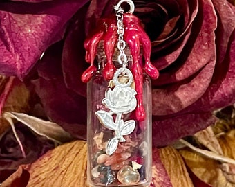 Puede incluir: Una pequeña botella de vidrio con un dije de rosa plateado y cera roja goteando por los lados. La botella está llena de varios cristales y piedras, incluyendo cuarzo claro, cuarzo rosa y amatista.
