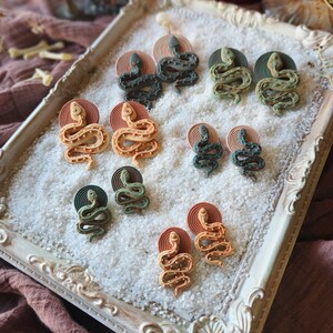 Op de afbeelding: Een set van 10 slangen oorbellen in verschillende kleuren, waaronder groen, bruin en oranje. De oorbellen worden gepresenteerd op een wit dienblad met een decoratieve rand. De oorbellen zijn gemaakt van polymer clay en hebben een getextureerd oppervlak.