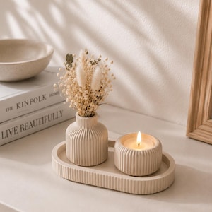 Peut inclure: Un plateau ovale beige à texture côtelée supportant un vase soliflore côtelé assorti rempli de fleurs séchées et un petit bougeoir côtelé contenant une bougie blanche allumée. L'ensemble présente une finition mate et des rainures verticales.