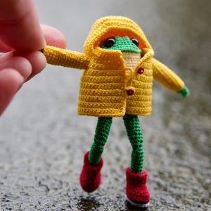 May include: A crocheted frog wearing a yellow raincoat and red boots. The frog is standing on a wet, grey surface.