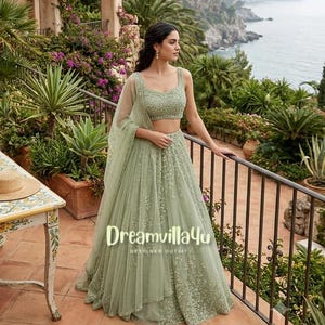 Op de afbeelding: Een saliegroene lehenga-set bestaande uit een mouwloze crop top en een rok tot op de grond, beide versierd met gedetailleerd borduurwerk en kralen in dezelfde kleur. Een bijpassende transparante dupatta hangt over één schouder.
