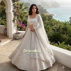 May include: A white lehenga choli set featuring intricate silver embroidery and beadwork throughout the skirt and blouse. The ensemble includes a sheer, lightweight dupatta draped over the shoulder, with the text Dreamvilla4u visible on the skirt.