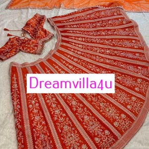 Può includere: Una gonna lehenga ricamata in rosso e bianco con una blusa abbinata ricamata in rosso e bianco. La gonna è realizzata in un tessuto fluido e ha una silhouette svasata. La blusa ha una scollatura a cuore e maniche corte. La gonna e la blusa sono decorate con intricati motivi floreali e paillettes. La gonna ha un orlo a balze. Il testo "Dreamvilla4u" è visibile sulla gonna.
