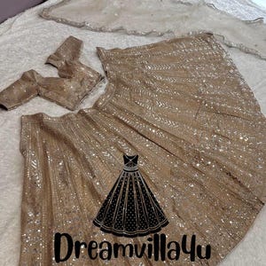 Könnte beinhalten: Ein goldfarbener und silberner Paillettenrock mit einer passenden bestickten Bluse. Der Rock hat ein fließendes Design und ist mit Pailletten und Perlen verziert. Die Bluse hat einen V-Ausschnitt und ist ebenfalls mit Pailletten und Perlen verziert. Der Text "Dreamvilla4u Designer Outfit" ist auf dem Rock aufgedruckt.