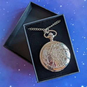 May include: A silver pocket watch with detailed engraved patterns, displayed in an open black box. The watch has a chain and a decorative face. The box's interior is dark, contrasting with the watch's shine.