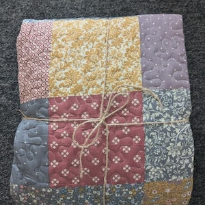 May include: A folded patchwork quilt featuring a variety of fabric squares in shades of mustard yellow, dusty rose, slate blue, and lavender. The quilt has a quilted texture with visible stitching and is tied together with a thin brown twine string.