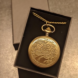 May include: A gold-coloured pocket watch with detailed engraved patterns, displayed in a black box. The watch has a chain and a complex face with circular designs. The box is open, showing the watch against a dark interior.