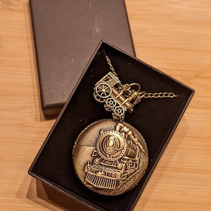 May include: A bronze-toned pocket watch with a train motif, displayed in a black box. The watch showcases a detailed engraving of a steam train on its face and hangs from a matching chain. A vintage-style accessory.