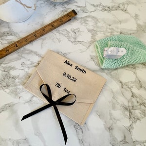 May include: A cream fabric envelope tied with a black ribbon, with the name "Alba Smith" and the date "9.10.22" printed in black. The envelope also indicates a weight of "7lb 1oz". A light green knitted baby hat is also in the image.