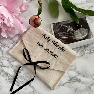 May include: A white fabric envelope with a black ribbon tied around it. The envelope is embroidered with the text "Baby Murphy Due 20.10.22".