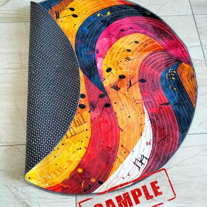 May include: A colorful round rug with a musical note design. The rug has a gray backing and is rolled up. The rug has a red stamp that says "SAMPLE".