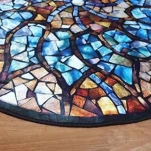 May include: A round rug with a stained glass design featuring blue, brown, and gold colours. The rug has a black border.