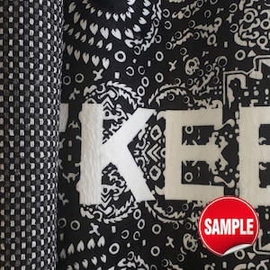 Puede incluir: Un tejido en blanco y negro con un patr&oacute;n de cachemira repetitivo y grandes letras blancas que forman parte de la palabra KEEP. Una pegatina circular roja con el texto blanco SAMPLE est&aacute; situada en la esquina inferior derecha de la tela.