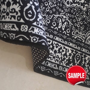 Puede incluir: Una alfombra en blanco y negro con un patr&oacute;n geom&eacute;trico y floral repetitivo. La alfombra est&aacute; doblada en una esquina, revelando una parte inferior oscura y texturizada y un borde negro. Se ve una pegatina roja con el texto blanco SAMPLE.