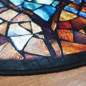 May include: A round rug with a stained glass design featuring a variety of colours, including blue, red, orange, and yellow. The rug has a black border with a stitched edge.