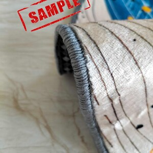 May include: Close-up of the edge of a rug with a gray border. The rug has a white background with a brown and white striped pattern.