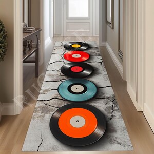 May include: A long, rectangular rug with a cracked concrete design. Five vinyl records are arranged vertically on the rug, each with a different colored center label. The records are black with a shiny finish.
