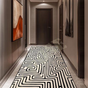 May include: A hallway runner rug with a black and cream-colored geometric maze pattern. The rug is rectangular and covers the length of the hallway. The walls are a light gray color with artwork and a dark brown door.