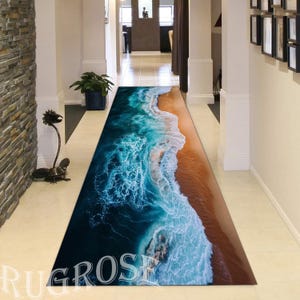 May include: A blue and white beach scene rug with a brown sandy beach and foamy waves. The rug is laid out in a hallway with white walls.