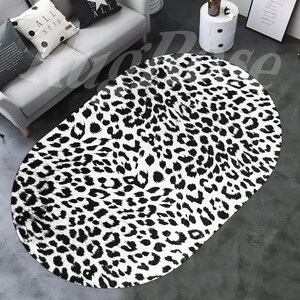 May include: Oval-shaped rug with a bold leopard print pattern in black and white. The rug is placed on a gray floor, and the design features irregular black spots against a white background. The rug's shape and pattern create a striking visual element.