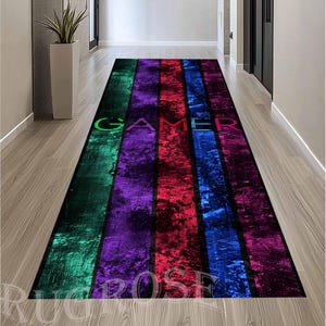 Può includere: Un tappeto colorato con un designo a strisce in tonalità di verde, viola, rosso, blu e rosa. La parola "GAMER" è stampata in bianco sul tappeto.