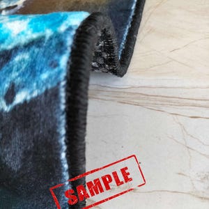 May include: Close-up of the edge of a blue and black rug with a black border. The rug has a galaxy-like pattern. The word "SAMPLE" is stamped in red on the rug.