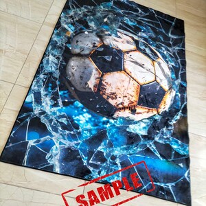 May include: A black and blue rug with a cracked glass design and a soccer ball in the center. The soccer ball is brown and white with black stitching. The rug has the word "SAMPLE" stamped in red on the bottom right corner.