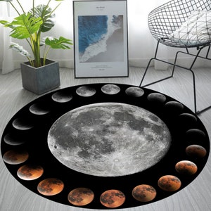May include: A round black and white rug featuring a large full moon in the centre surrounded by a ring of crescent moons in various phases. The rug has a black background with a white moon and a ring of orange crescent moons.