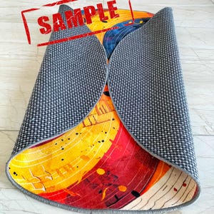 May include: A round rug with a colorful abstract design featuring yellow, red, and blue hues. The rug has a gray backing and is rolled up.