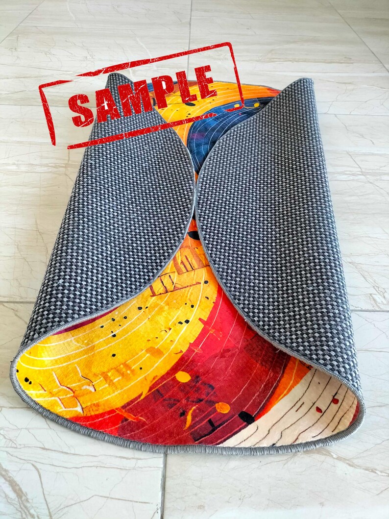 May include: A round rug with a colorful abstract design featuring yellow, red, and blue hues. The rug has a gray backing and is rolled up.