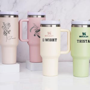 May include: Four stainless steel travel mugs with handles. The mugs are in different colors: pink, cream, and green. Each mug has a personalized name on it: June, Dwight, Trista. The mugs are on a white surface.