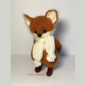 May include: A crocheted fox plush toy wearing a white scarf with a pom-pom. The fox has brown fur, black eyes, and a black nose.